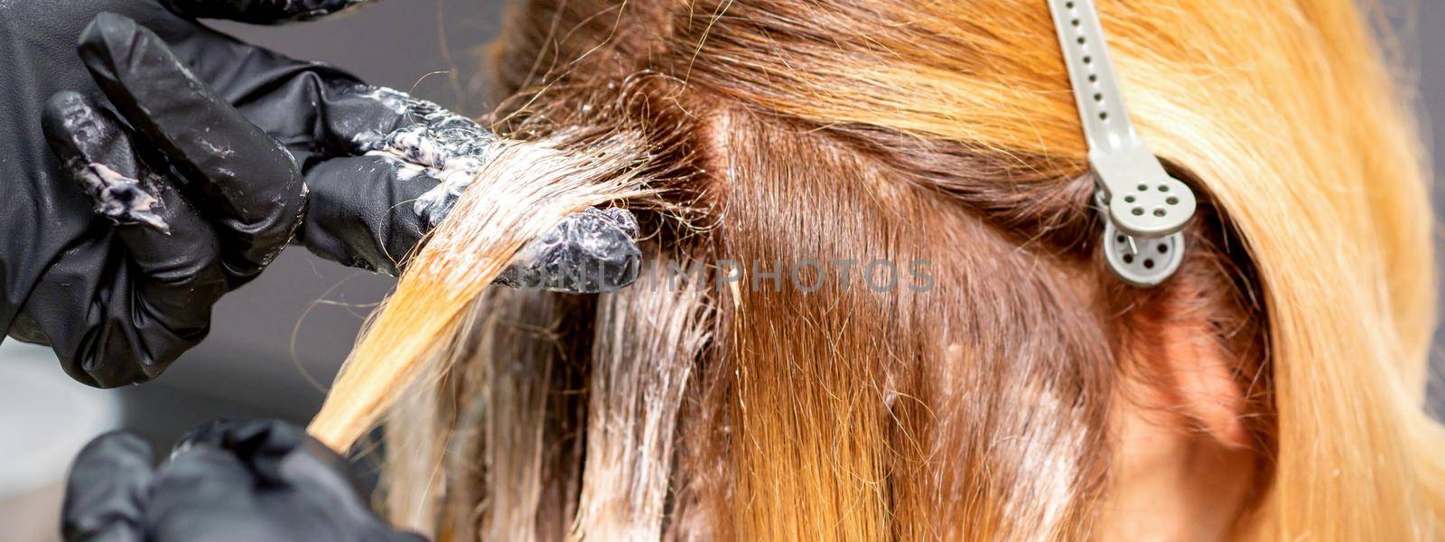 Hands of the hairdresser are applying the dye with gloved fingers on the red hair of a young red-haired woman in a hairdresser salon, close up