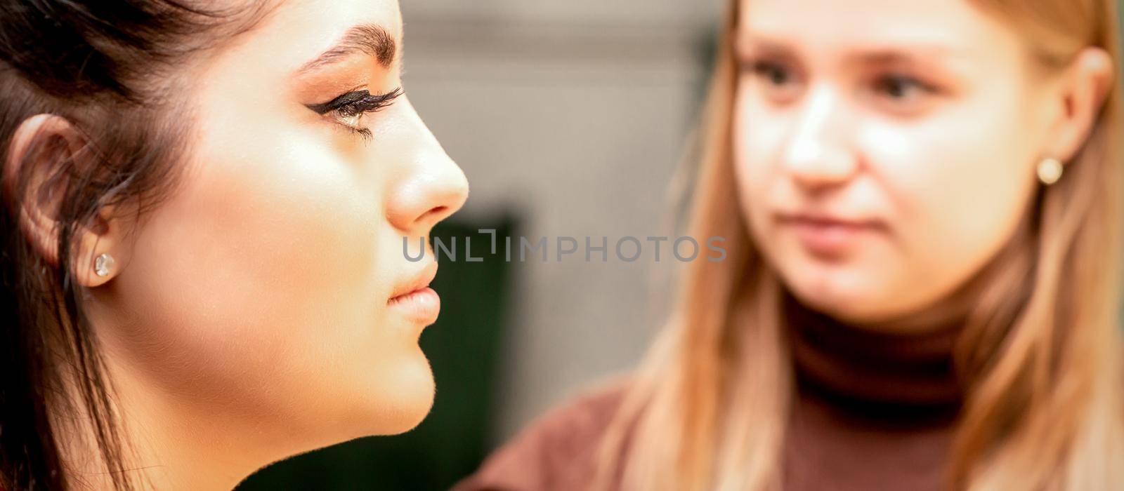 Profile portrait of the beautiful female model with long hair and makeup with a makeup artist in a beauty salon. by okskukuruza