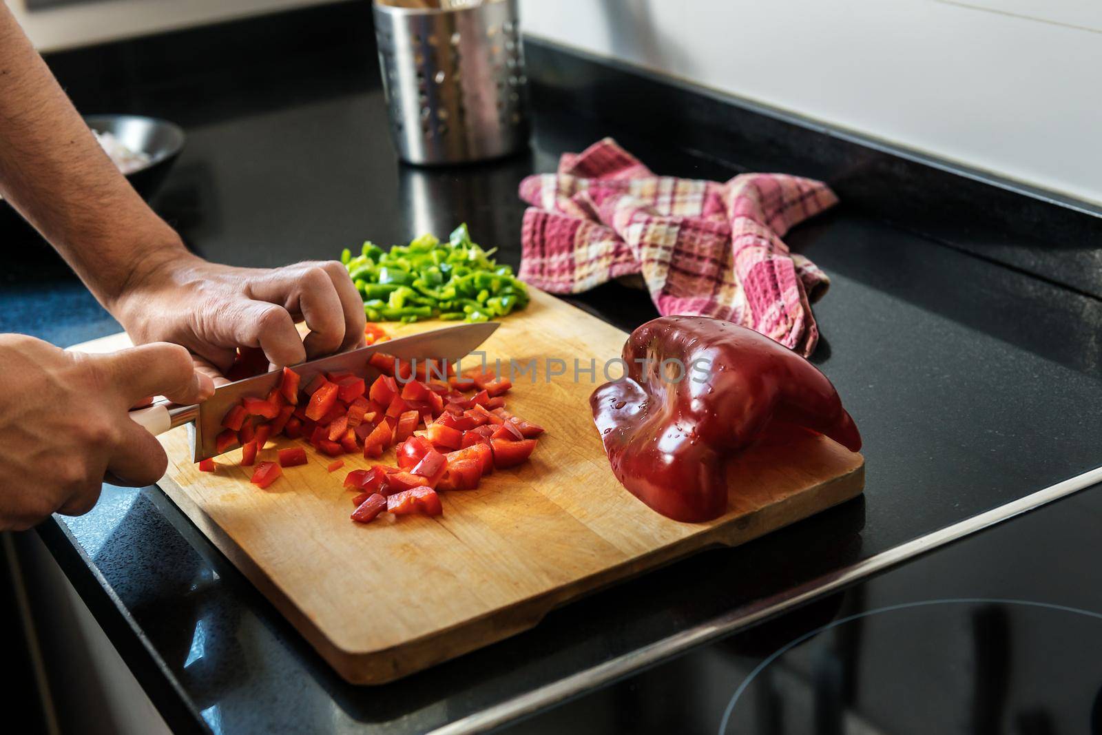 male hands chopping red pepper on a wooden board, in the background you see chopped green pepper and a kitchen rag