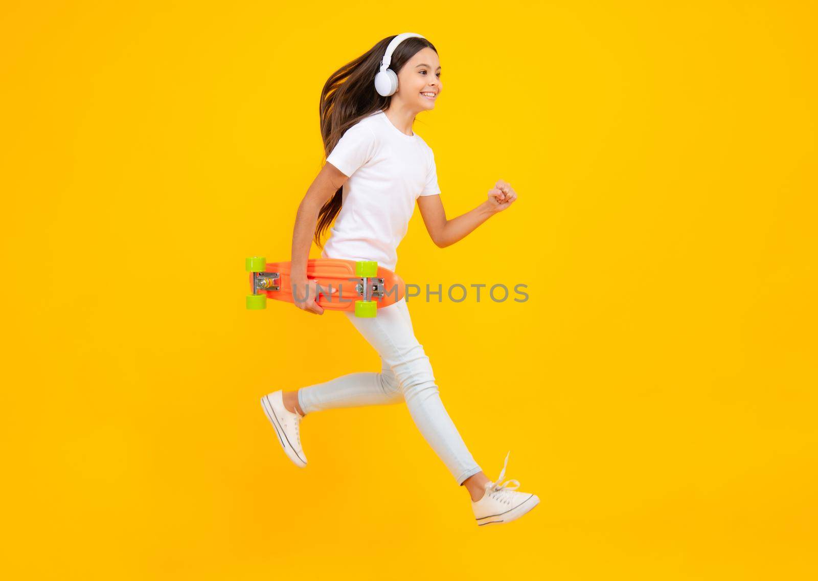 Happy teenager portrait. Jump and run. Fashion teenage lifestyle, beautiful teen girl with skateboard and headphones isolated on studio background. Smiling girl