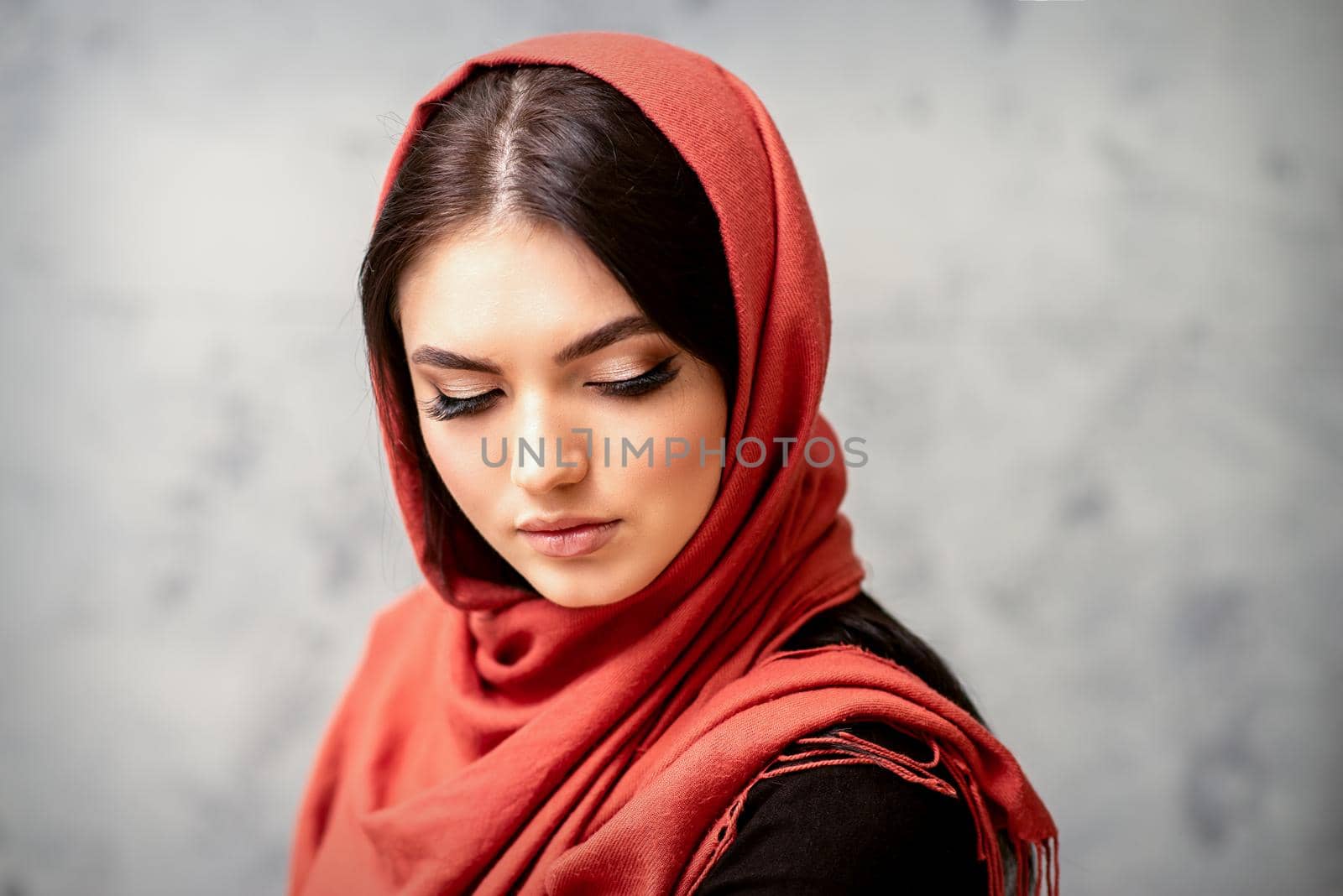 The fashionable young woman. Portrait of the beautiful female model with long hair and makeup with eyelash extensions in a red scarf. Beauty young caucasian woman on the background of a gray wall