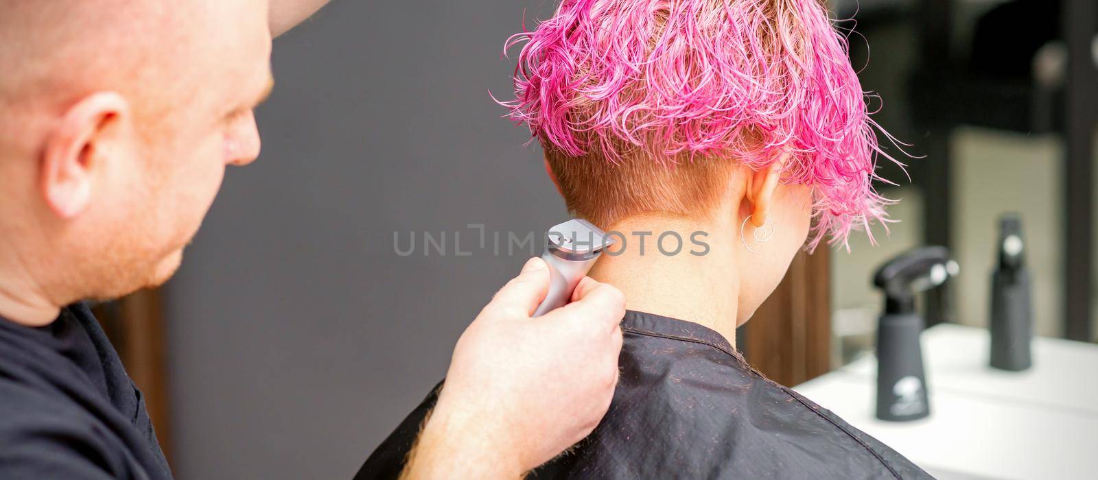 Male hairdresser shaves neck of a young caucasian woman with a short pink hairstyle by electric shaver in a hairdresser salon, close up
