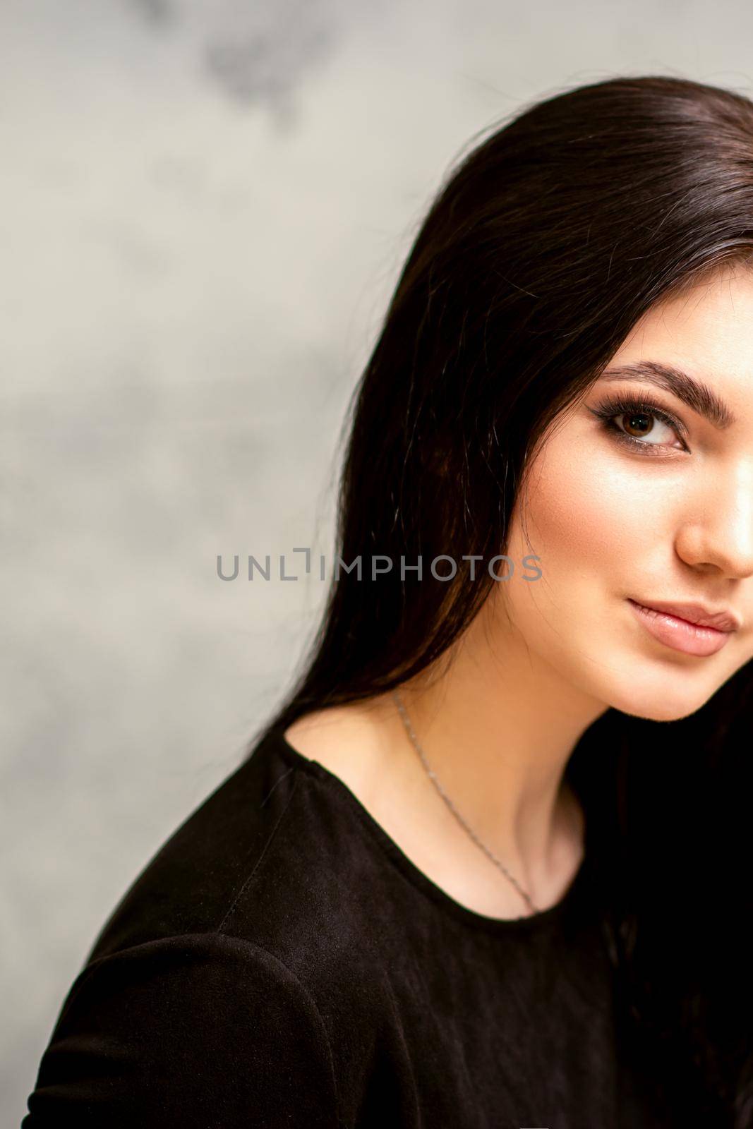 The fashionable young woman. Portrait of the beautiful female model with long hair and makeup. Beauty young caucasian woman with a black hairstyle on the background of a gray wall