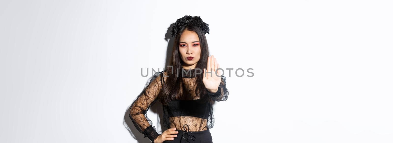 Image of serious asian woman in halloween costume of witch, showing stop gesture, forbid or prohibit something with displeased confident face, standing white background by Benzoix