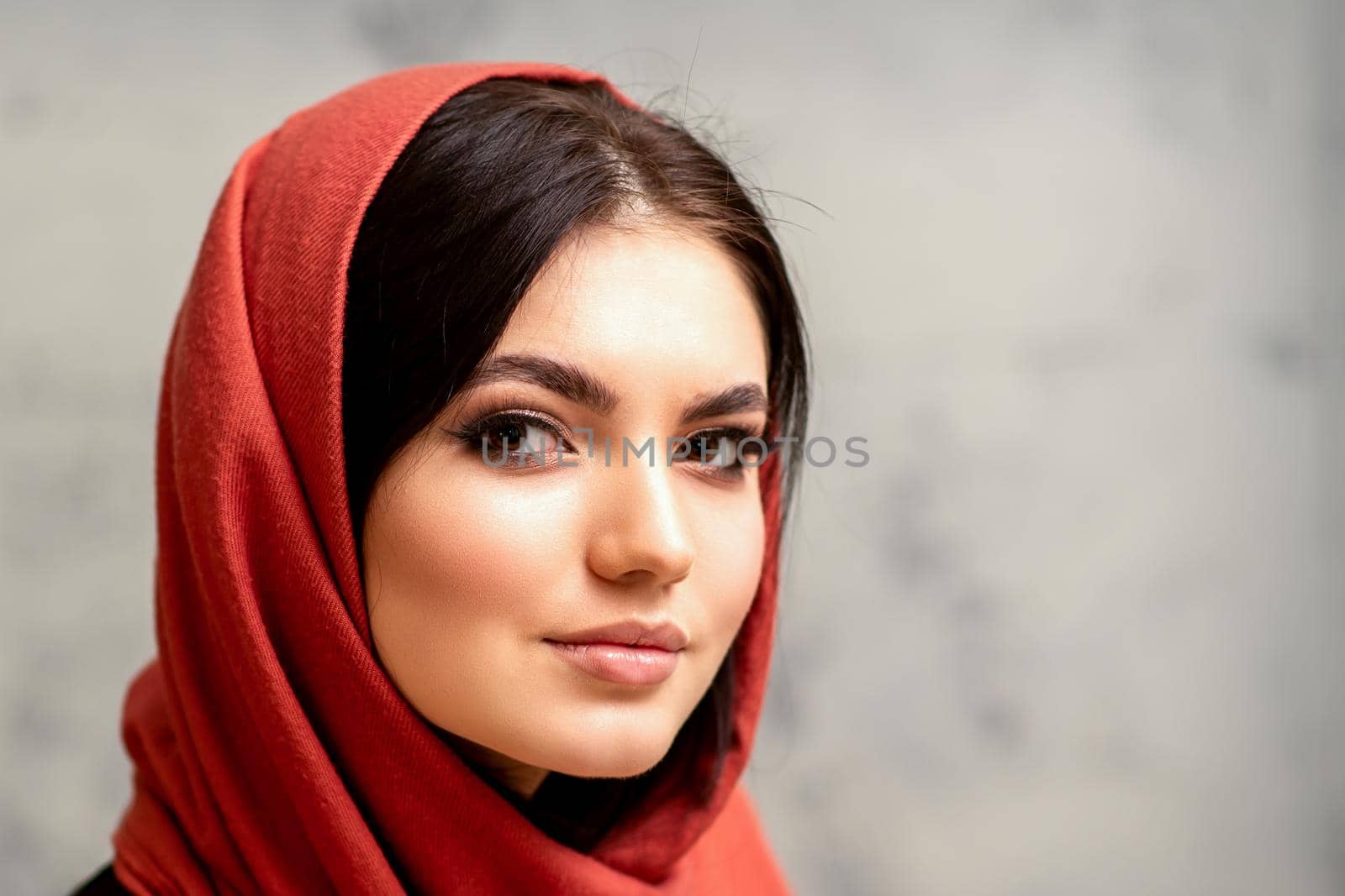 The fashionable young woman. Portrait of the beautiful female model with long hair and makeup in a red scarf. Beauty young caucasian woman on the background of a gray wall