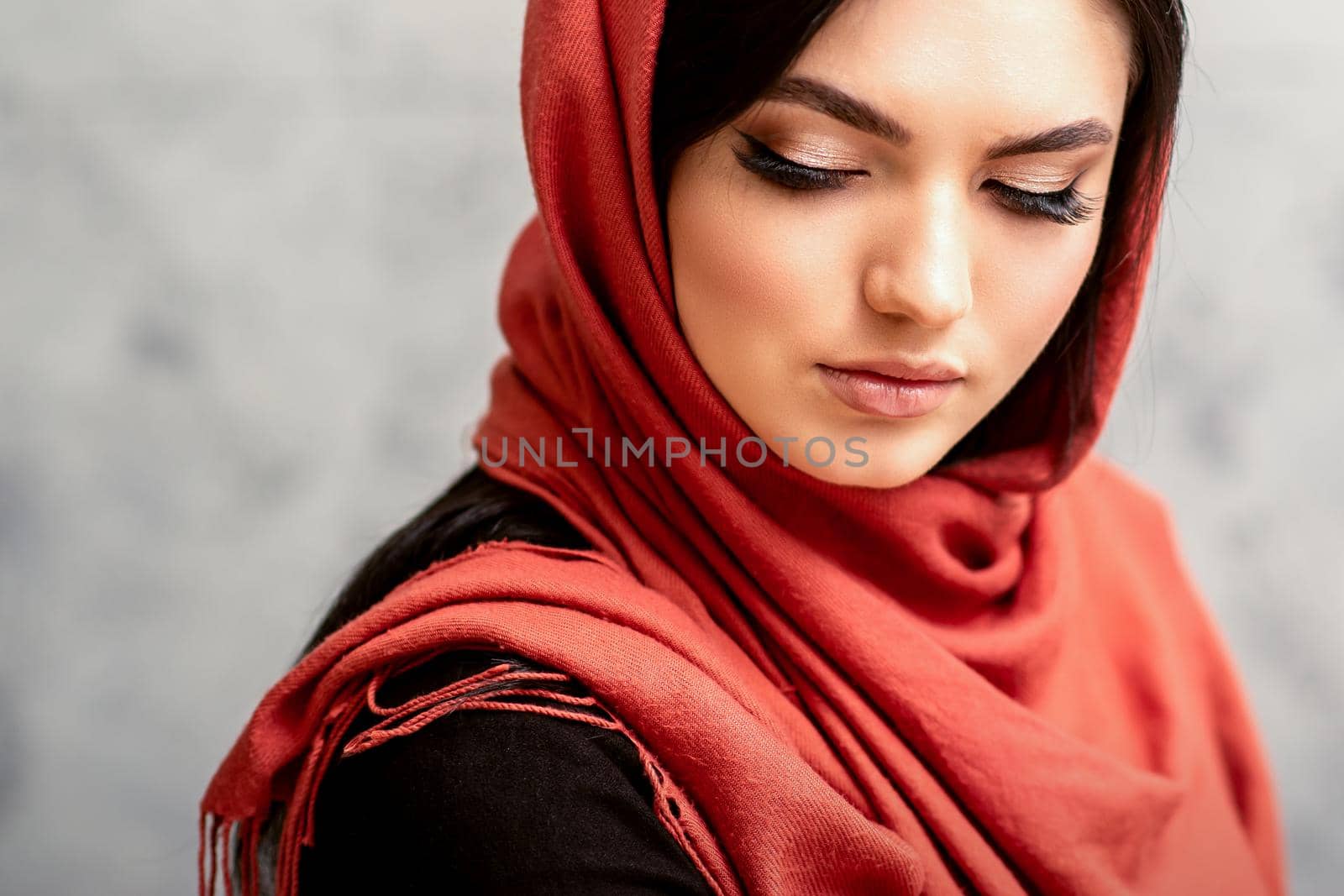 The fashionable young woman. Portrait of the beautiful female model with long hair and makeup with eyelash extensions in a red scarf. Beauty young caucasian woman on the background of a gray wall