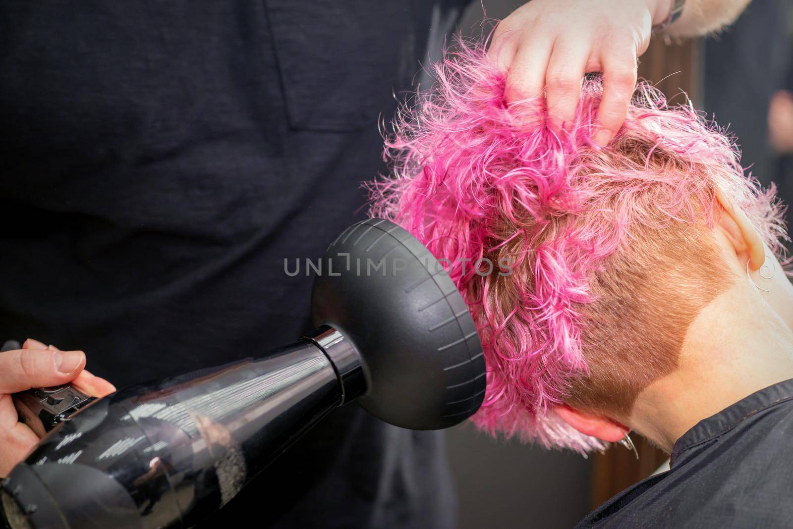 Drying short pink bob hairstyle of a young caucasian woman with a black hair dryer with the brush by hands of a male hairdresser in a hair salon, close up