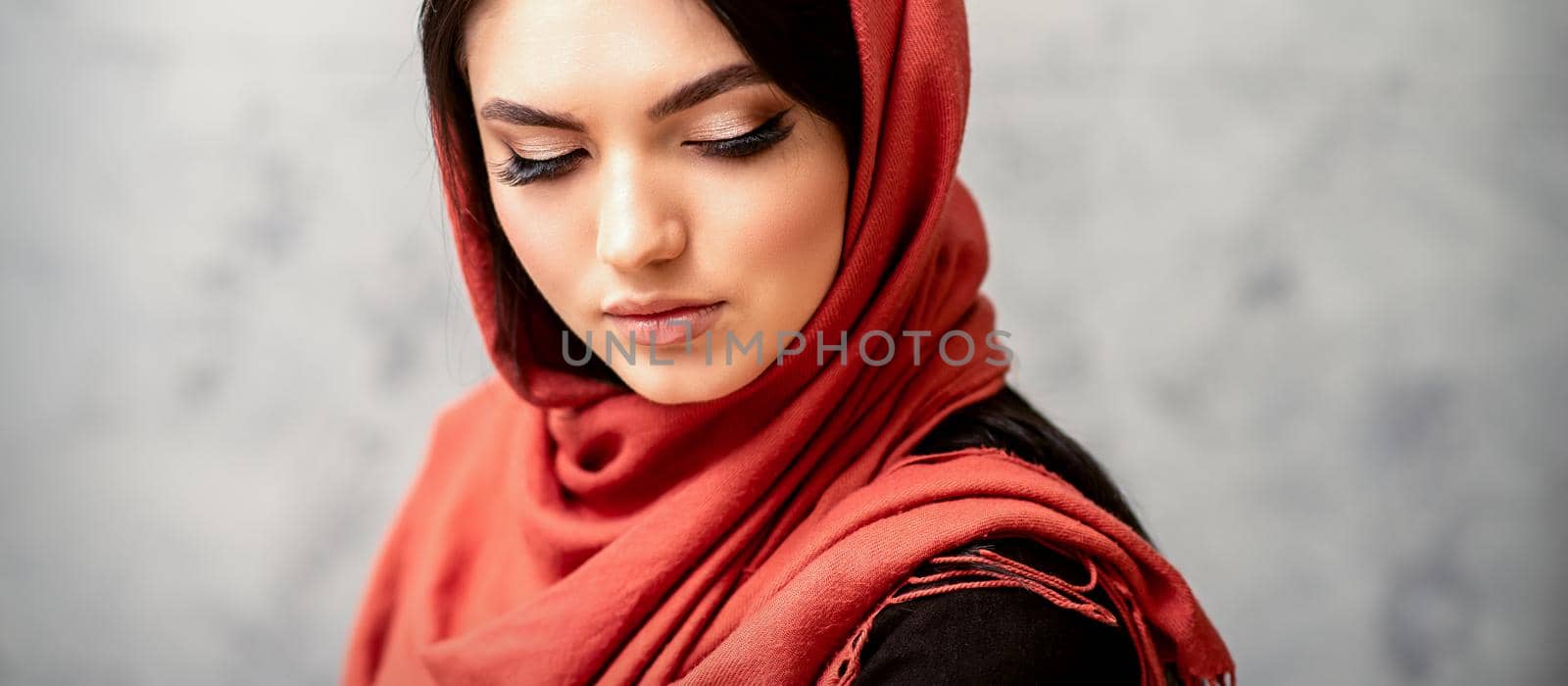 The fashionable young woman. Portrait of the beautiful female model with long hair and makeup with eyelash extensions in a red scarf. Beauty young caucasian woman on the background of a gray wall