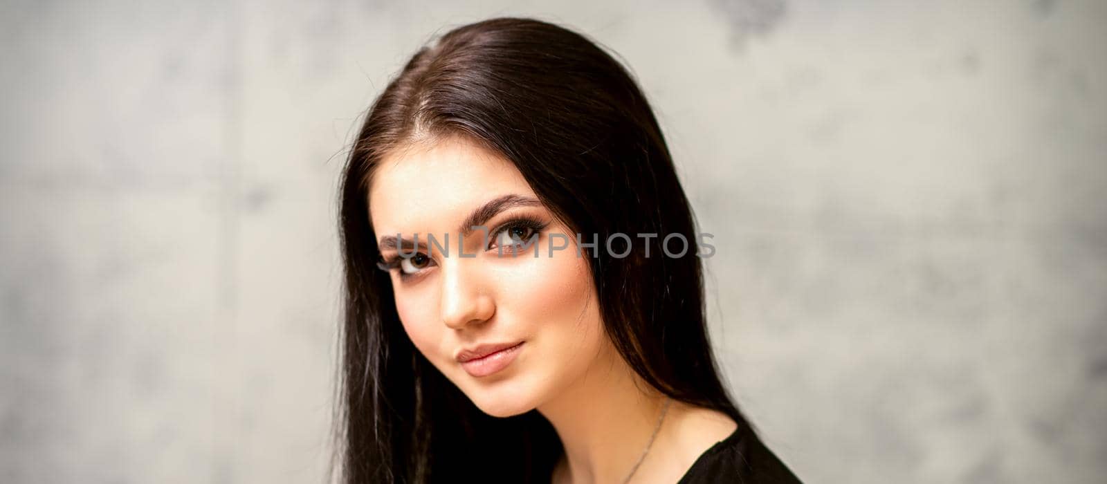 The fashionable young woman. Portrait of the beautiful female model with long hair and makeup. Beauty young caucasian woman with a black hairstyle on the background of a gray wall