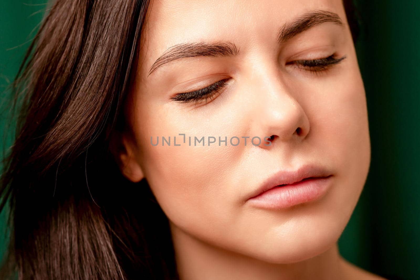 The fashionable young woman. Portrait of the beautiful female model with long hair and makeup with closed eyes. Beauty young woman with a brown curly hairstyle on the background of a green curtain