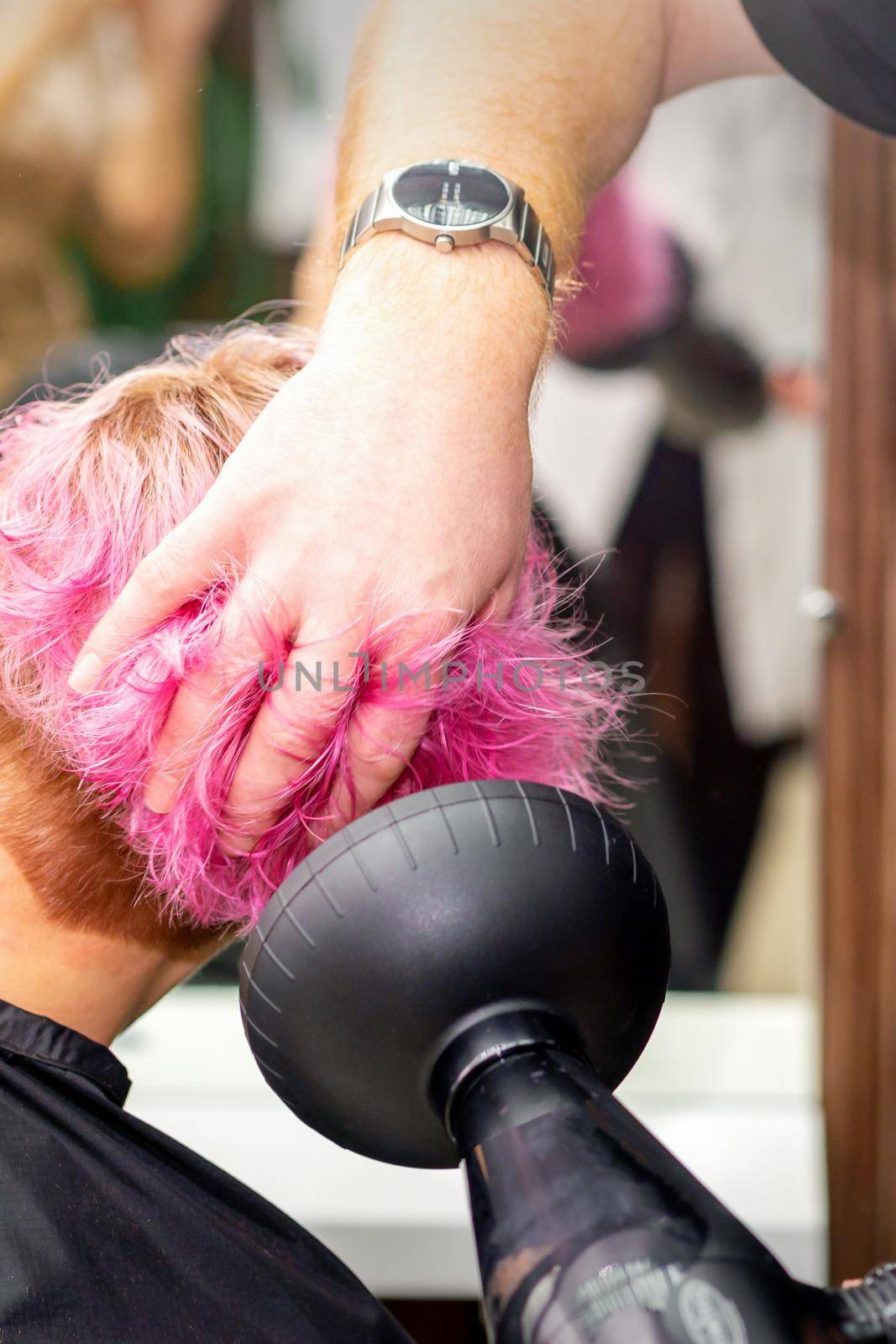 Drying short pink bob hairstyle of a young caucasian woman with a black hair dryer with the brush by hands of a male hairdresser in a hair salon, close up