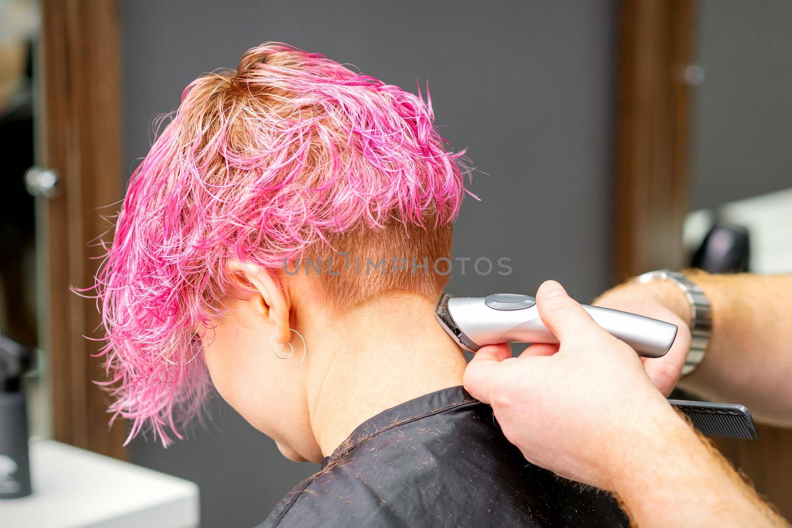 Male hairdresser shaves neck of a young caucasian woman with a short pink hairstyle by electric shaver in a hairdresser salon, close up