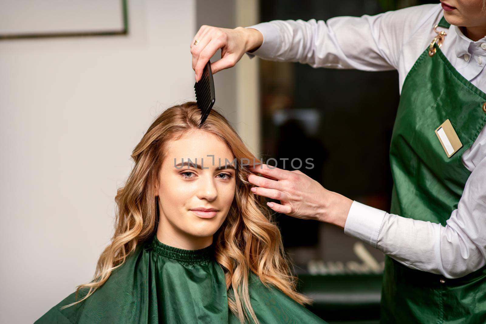 Hairdresser combing long hair of young caucasian woman looking at the camera and smiling in beauty salon. by okskukuruza