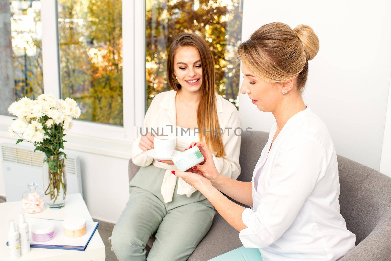 Beautician offering product for the young woman holding a white plastic jar with a cream sitting on the sofa