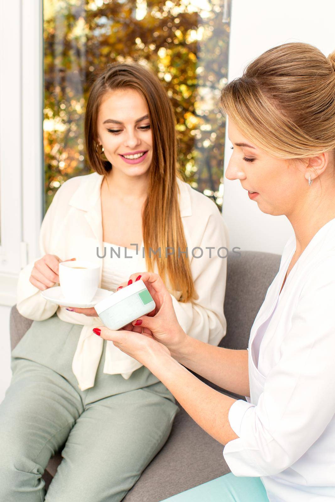 Beautician offering product for the young woman holding a white plastic jar with a cream sitting on the sofa