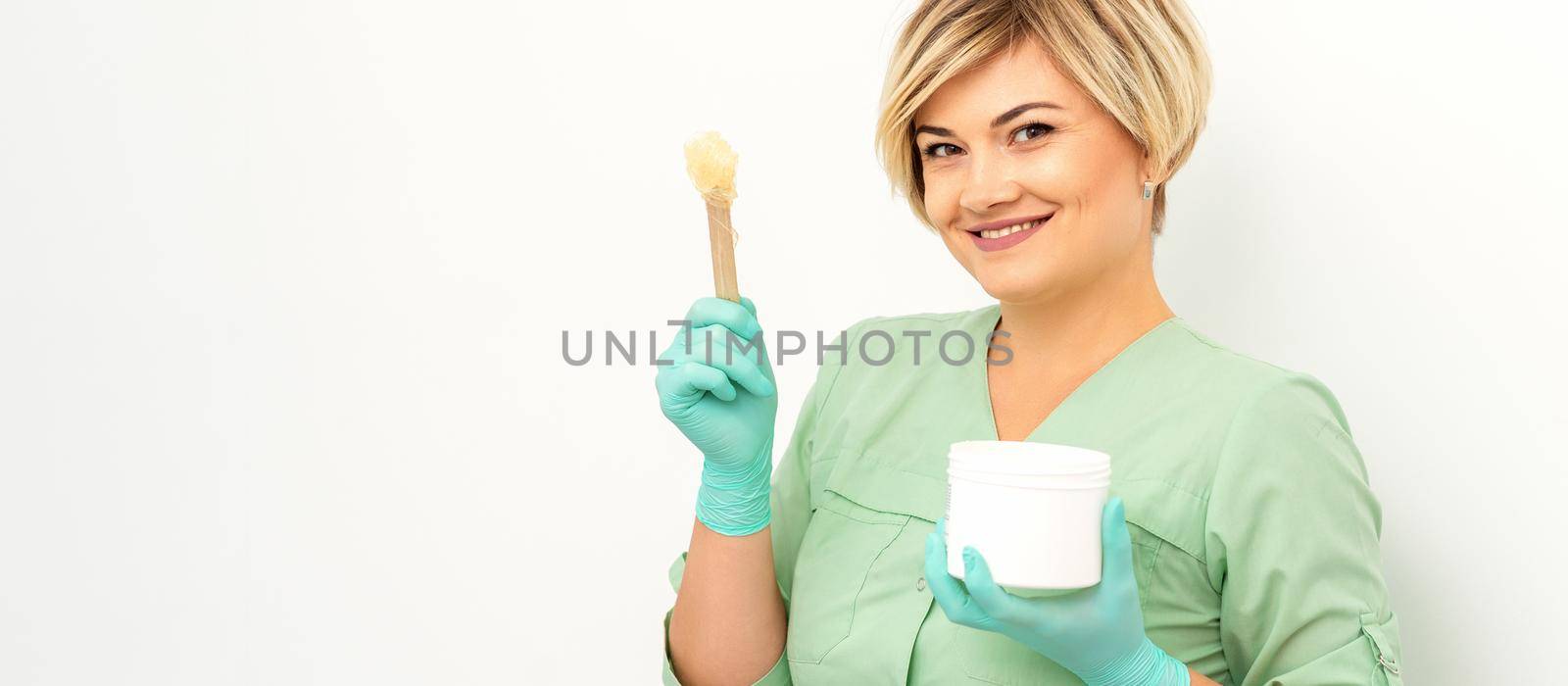 The master of sugar hair removal holds liquid yellow sugar paste, wax for depilation with the jar on a wooden stick on a white background