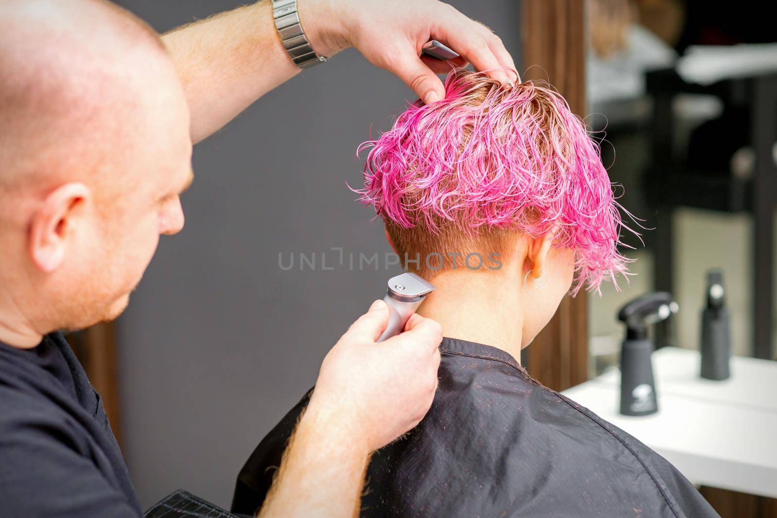 Male hairdresser shaves neck of a young caucasian woman with a short pink hairstyle by electric shaver in a hairdresser salon, close up. by okskukuruza