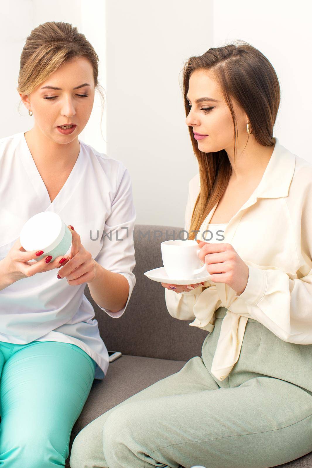 Beautician offering product for the young woman holding a white plastic jar with a cream sitting on the sofa