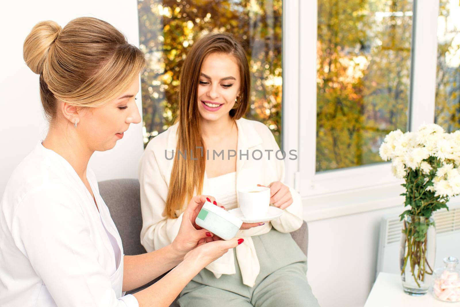 Beautician offering product for the young woman holding a white plastic jar with a cream sitting on the sofa