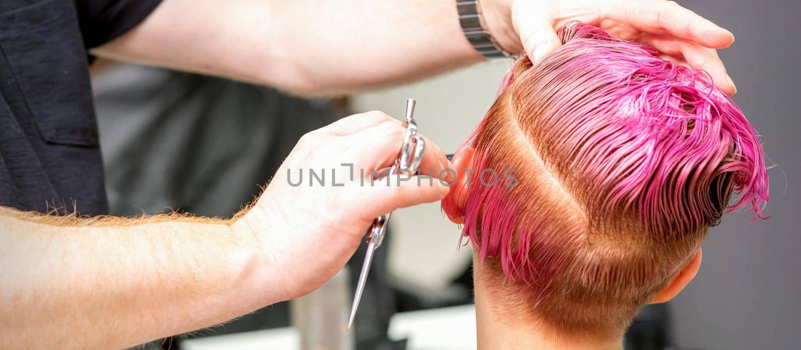 Haircut of dyed short pink wet hair of young caucasian woman by a male hairdresser in a barbershop