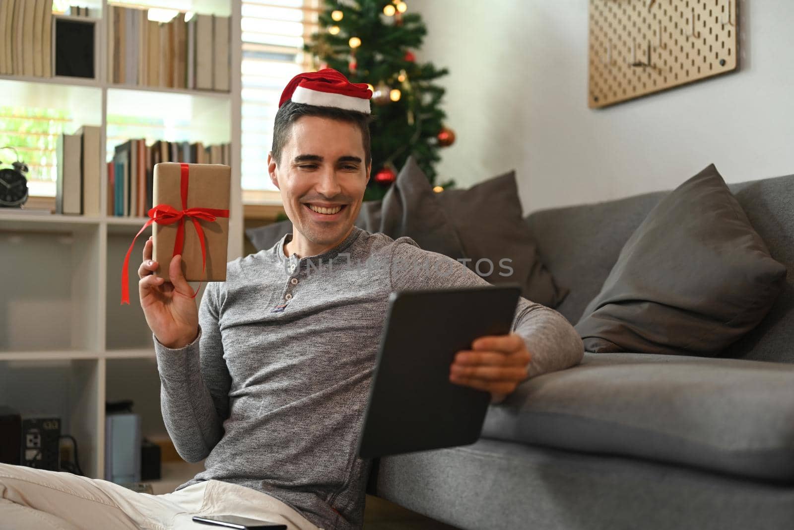 Smiling man wearing Santa hat showing Christmas gift box and talking with family or her friend on digital tablet.