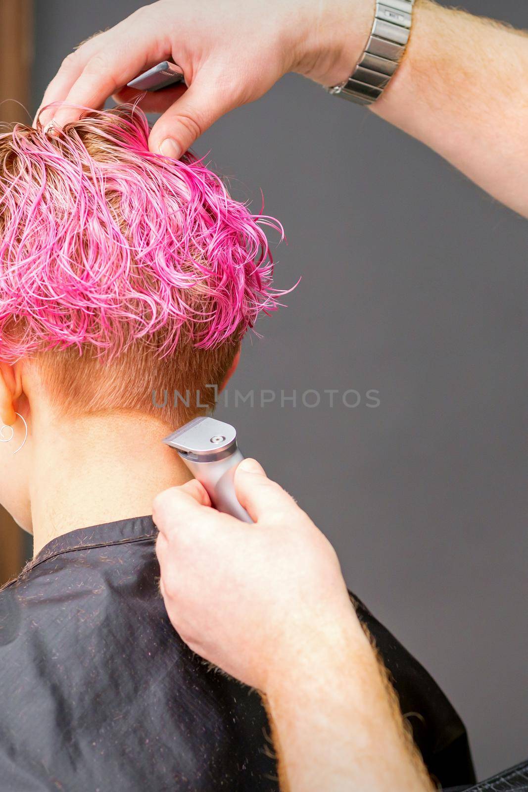 Male hairdresser shaves neck of a young caucasian woman with a short pink hairstyle by electric shaver in a hairdresser salon, close up. by okskukuruza