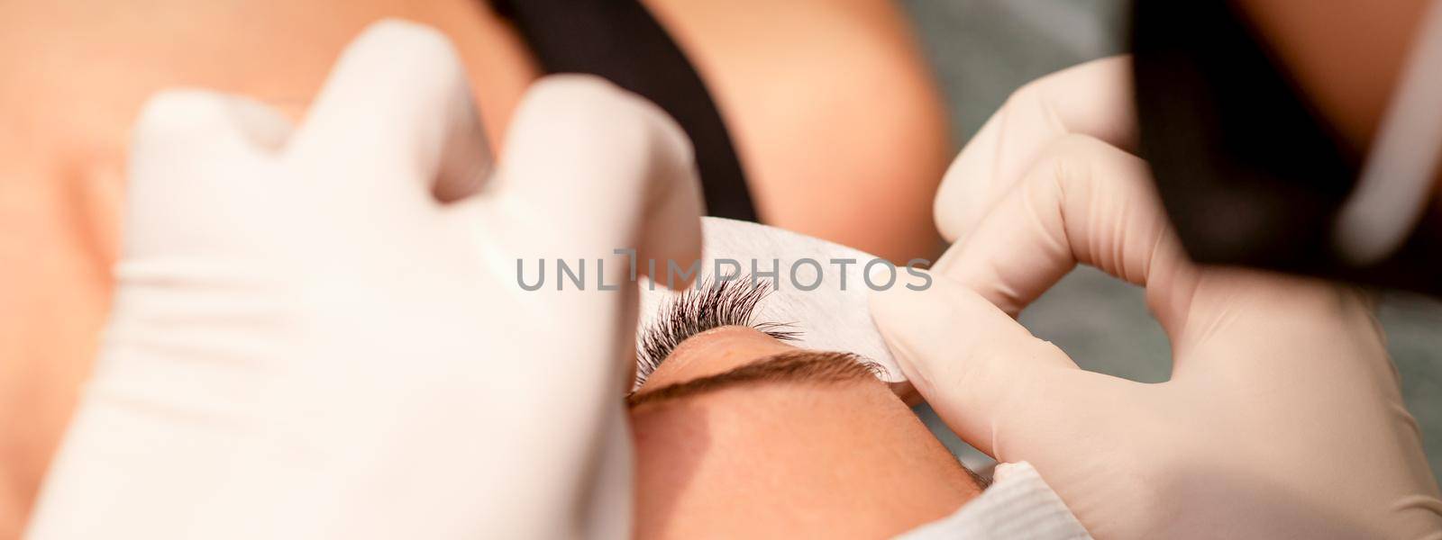 The hands of the cosmetologist are gluing white tape under the eye of the young caucasian woman during the eyelash extension procedure, closeup