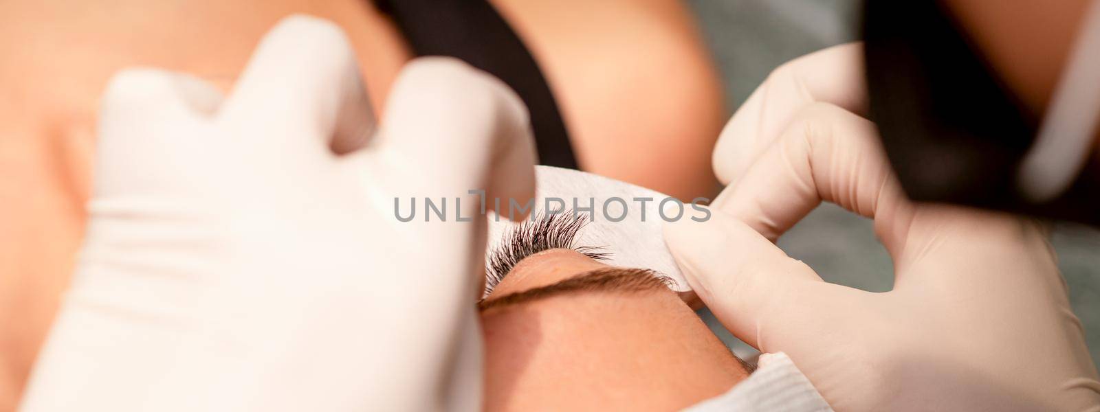 The hands of the cosmetologist are gluing white tape under the eye of the young caucasian woman during the eyelash extension procedure, closeup. by okskukuruza