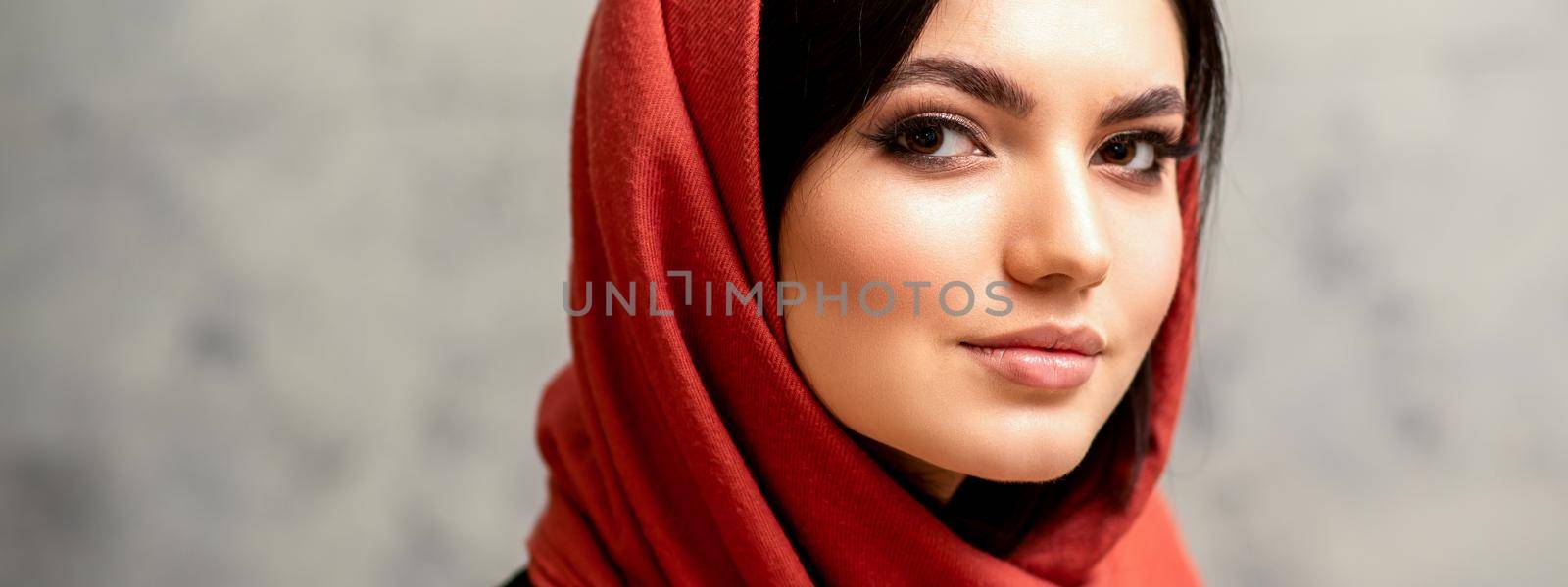 The fashionable young woman. Portrait of the beautiful female model with long hair and makeup in a red scarf. Beauty young caucasian woman on the background of a gray wall