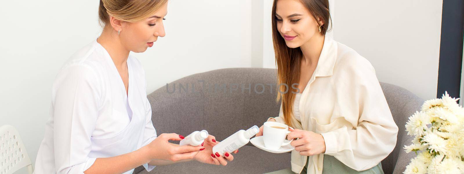 Beautician offering product for the young woman holding white plastic bottles with a cream sitting on the sofa