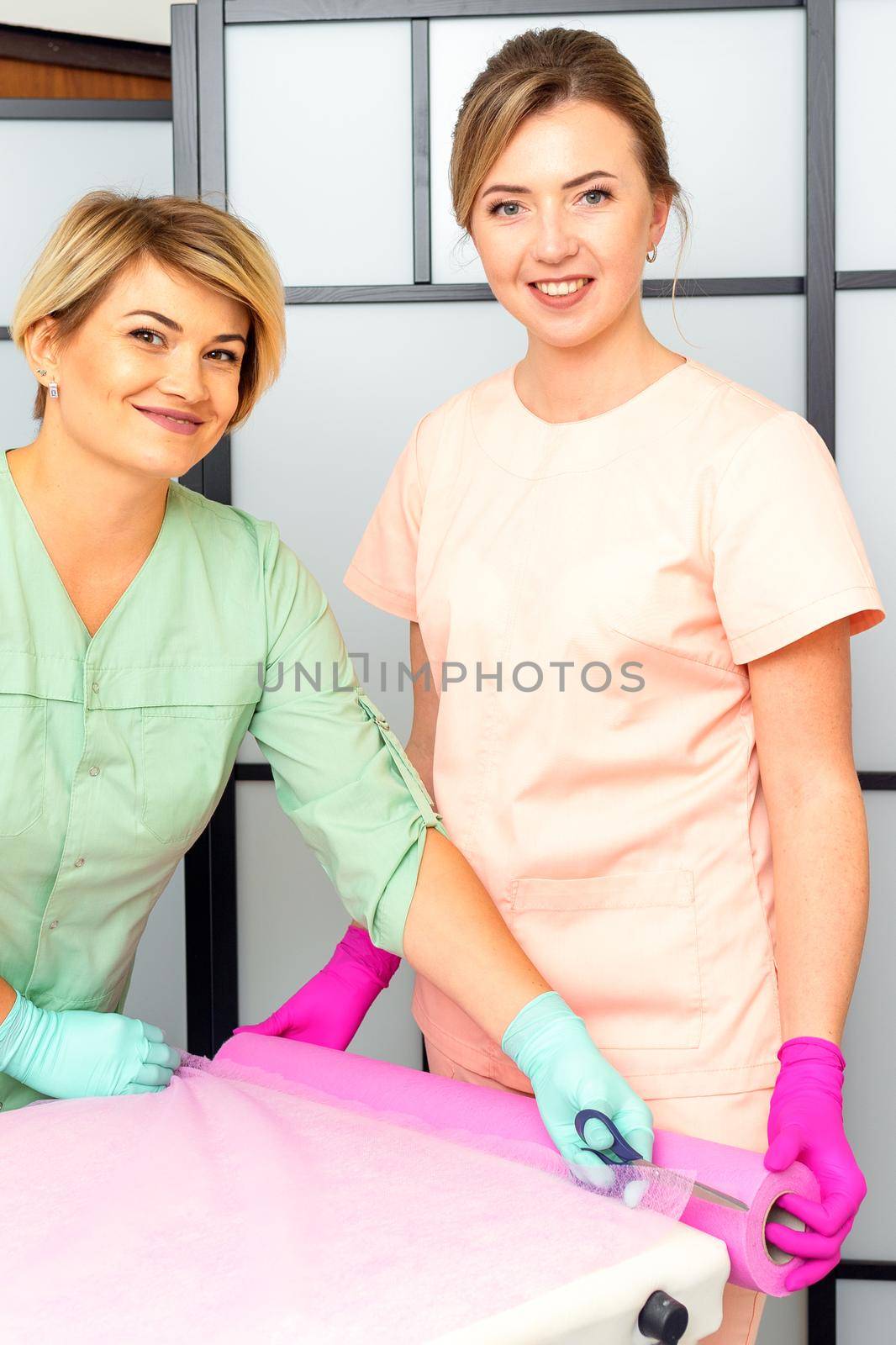 Two medical women cut disposable sheets with scissors during prepare the couch for the beauty procedure. by okskukuruza