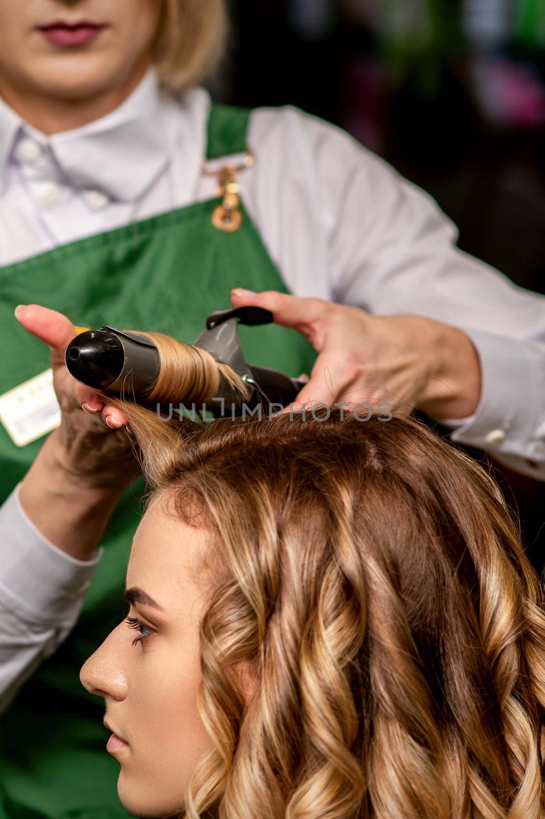 The female hairdresser is curling hair for a brown-haired young caucasian woman in a beauty salon