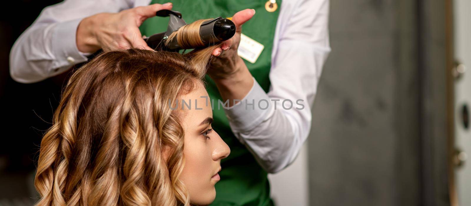 The female hairdresser is curling hair for a brown-haired young caucasian woman in a beauty salon