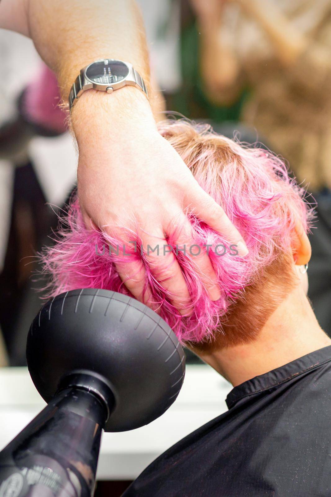 Drying short pink bob hairstyle of a young caucasian woman with a black hair dryer with the brush by hands of a male hairdresser in a hair salon, close up