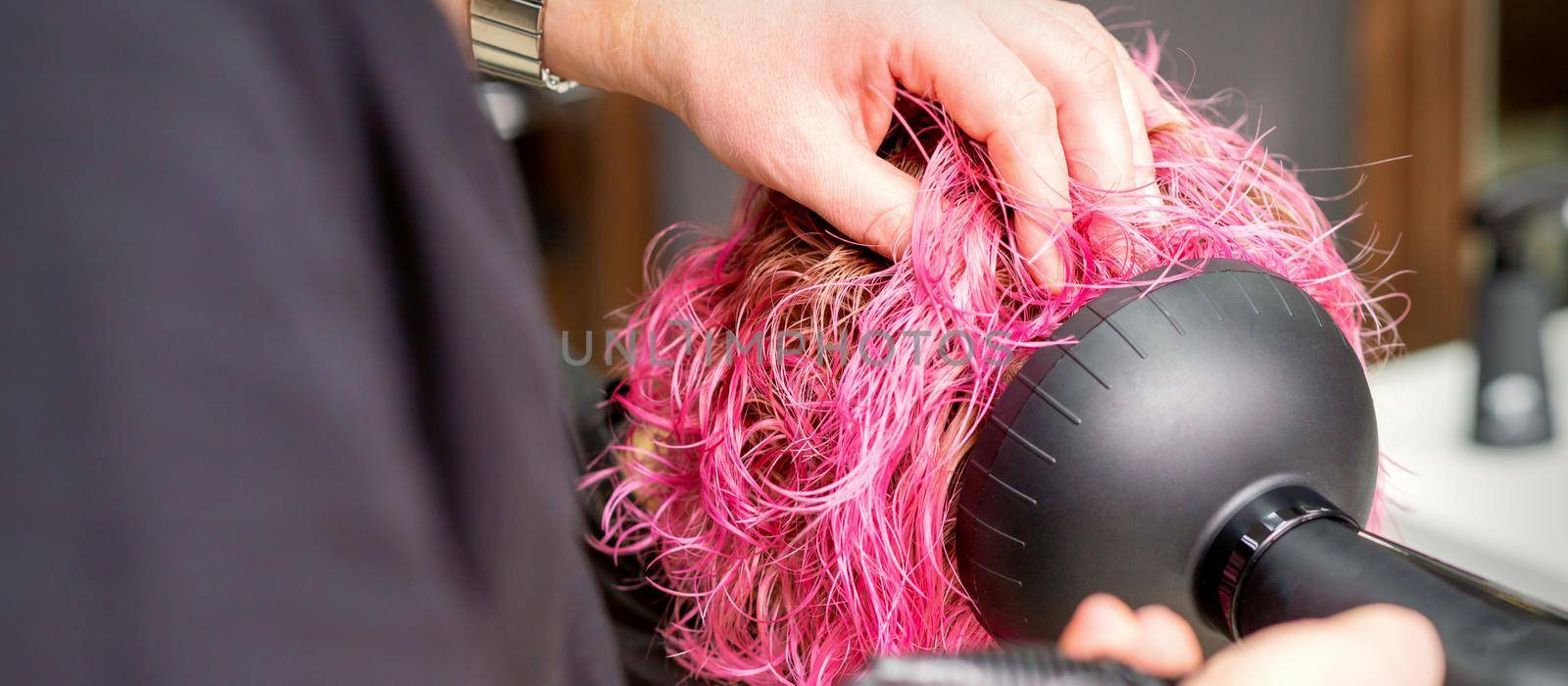 Drying short pink bob hairstyle of a young caucasian woman with a black hair dryer with the brush by hands of a male hairdresser in a hair salon, close up