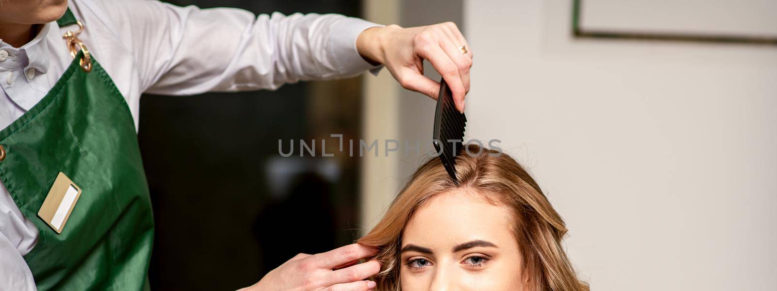 Hairdresser combing long hair of young caucasian woman looking at the camera and smiling in beauty salon. by okskukuruza