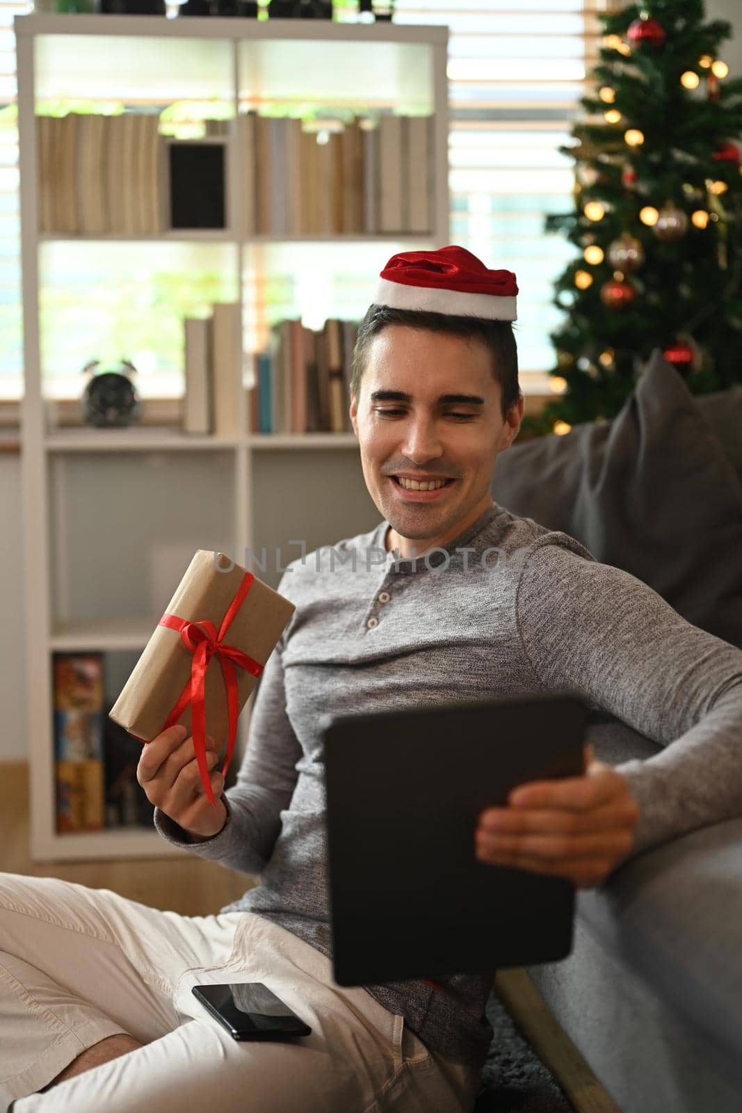 Young man wearing Santa hat holding Christmas gift box having video call with his family.