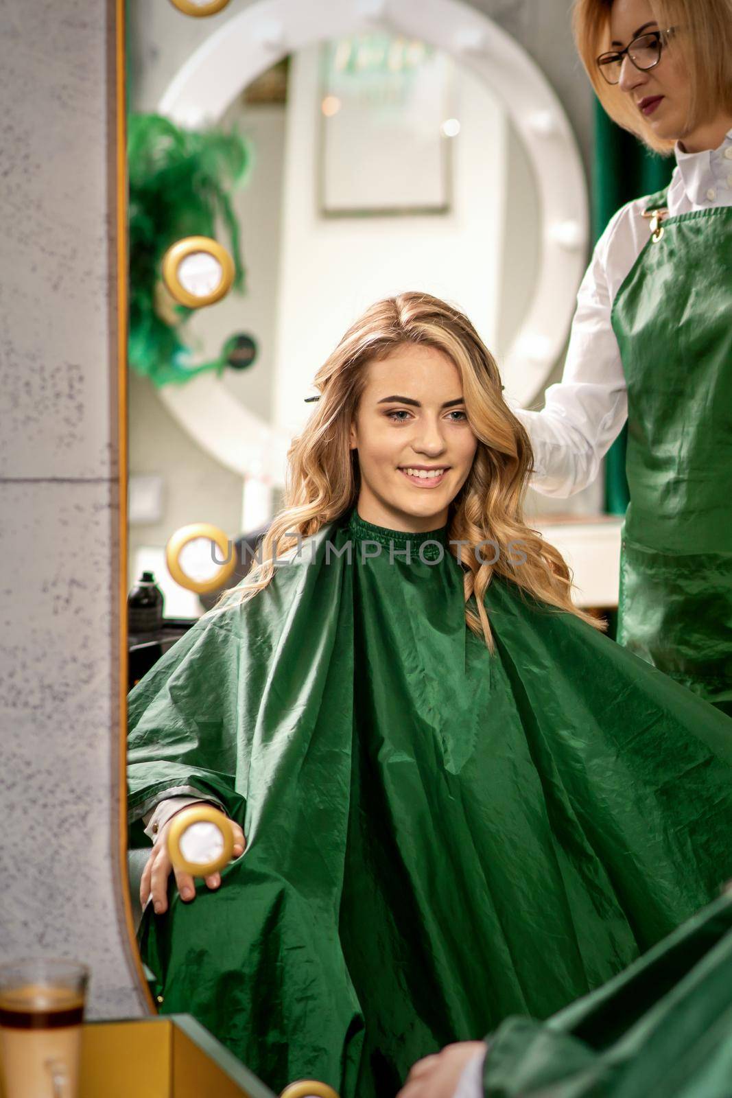 Hairdresser combing long hair of young caucasian woman looking and smiling in the mirror in beauty salon