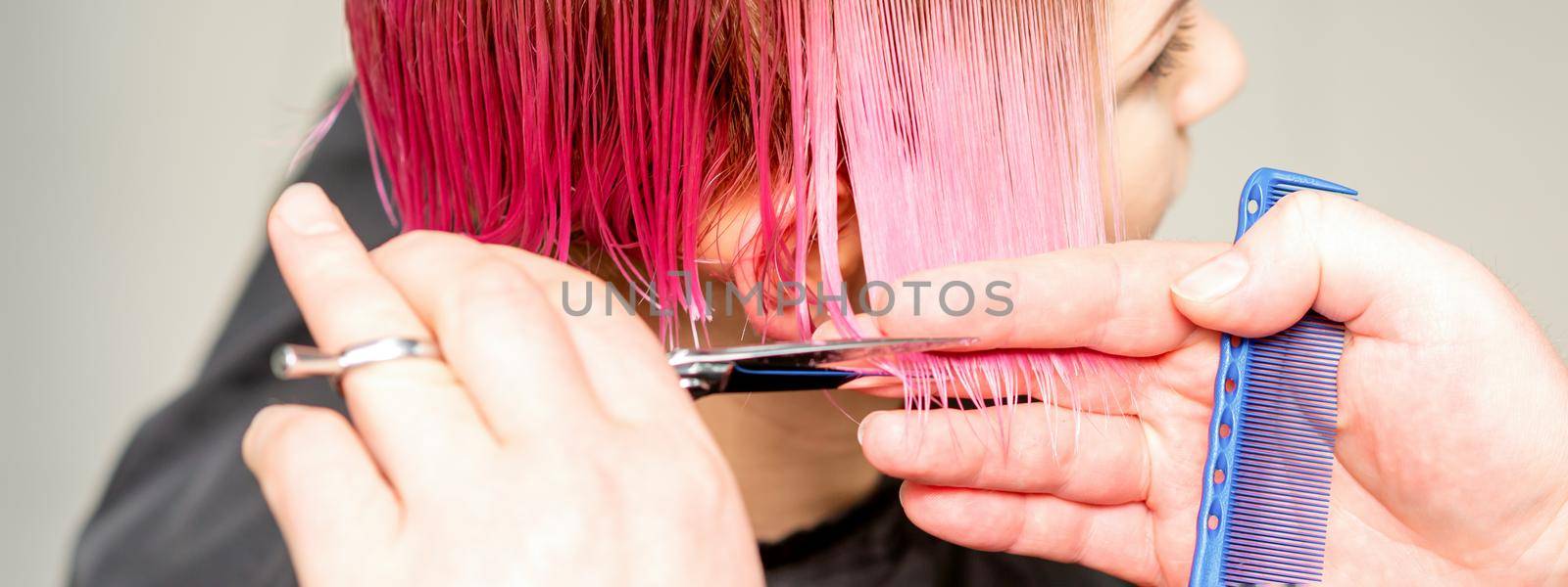 Haircut of dyed short pink wet hair of young caucasian woman by a male hairdresser in a barbershop