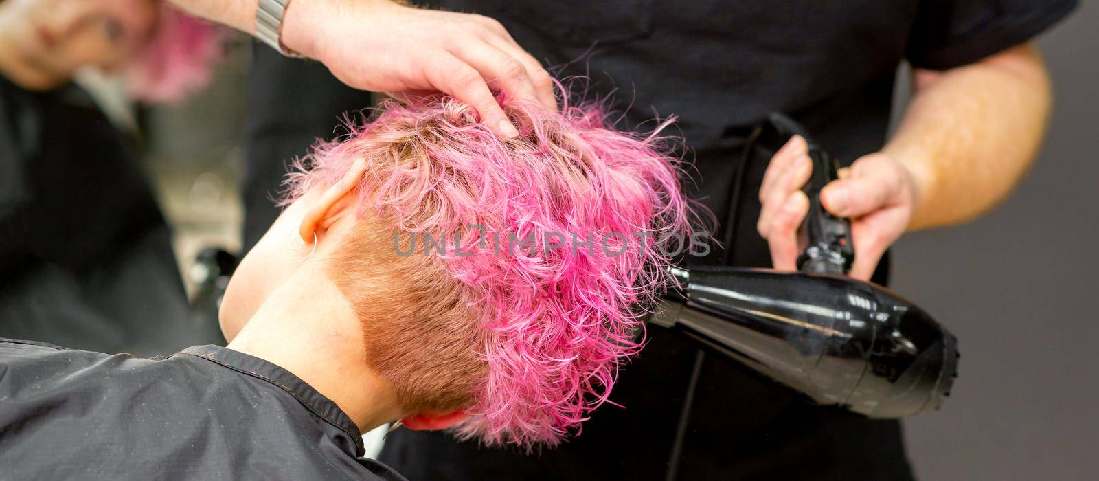 Drying short pink bob hairstyle of a young caucasian woman with a black hair dryer with the brush by hands of a male hairdresser in a hair salon, close up