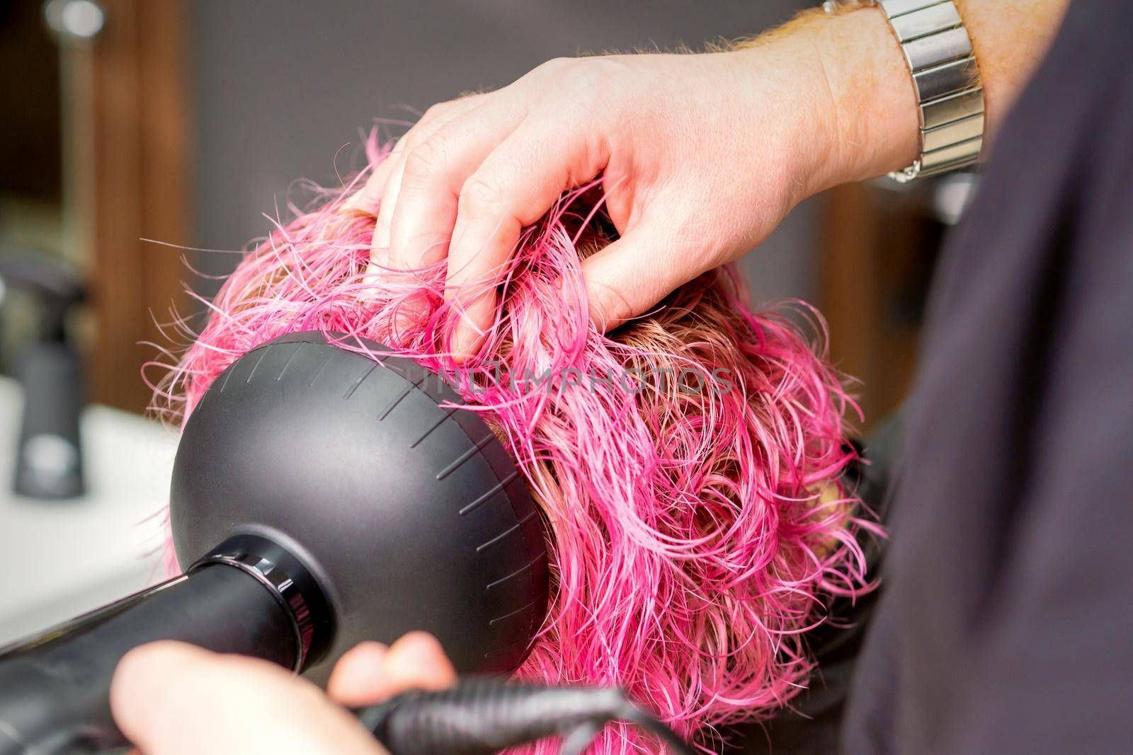 Drying short pink bob hairstyle of a young caucasian woman with a black hair dryer with the brush by hands of a male hairdresser in a hair salon, close up