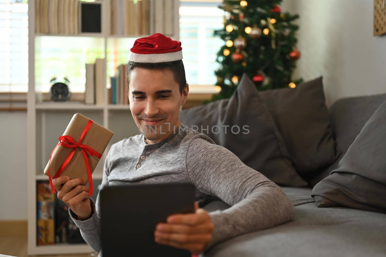 Man wearing Santa hat showing Christmas gift box and having video call with family or his friend on digital tablet.