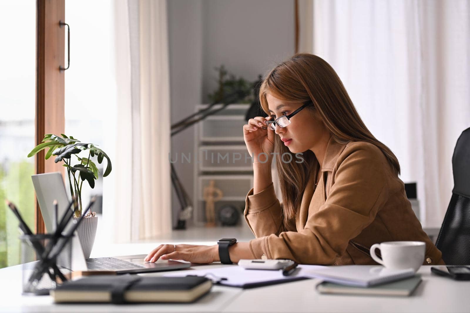 Young businesswoman holding her glasses and looking at laptop screen, working in internet at her office desk.