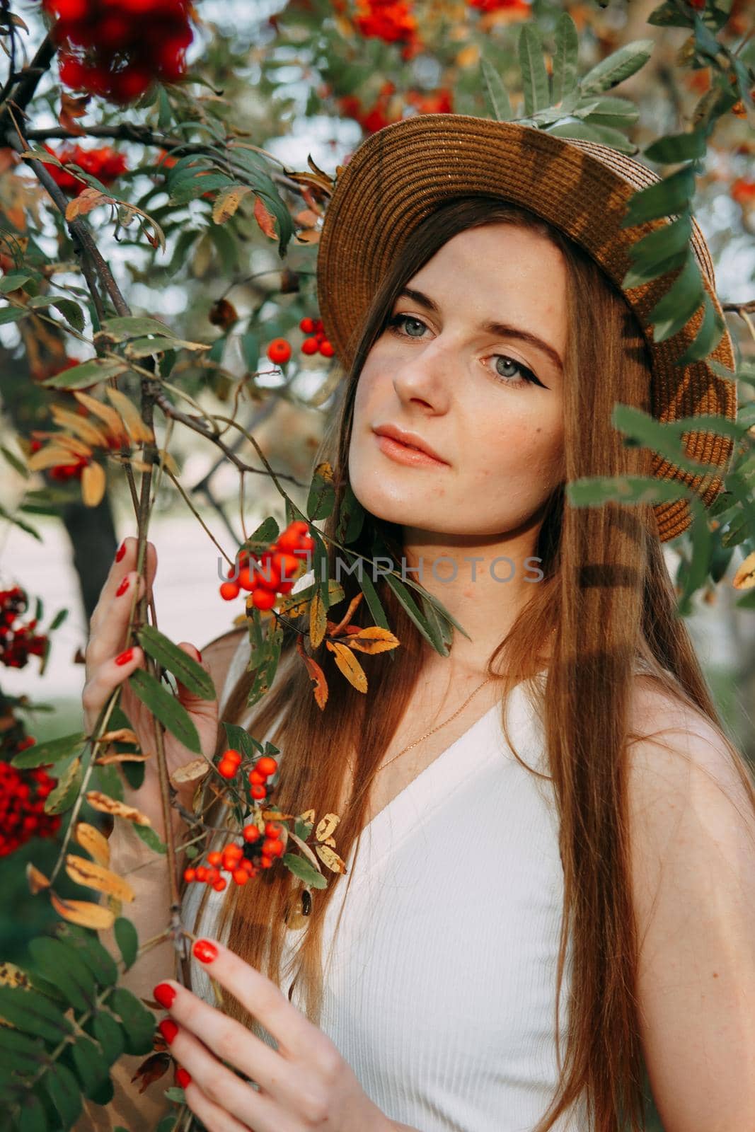 A beautiful blonde girl in a hat with long hair, on a walk in the autumn season. Portrait of a woman at a rowan tree.