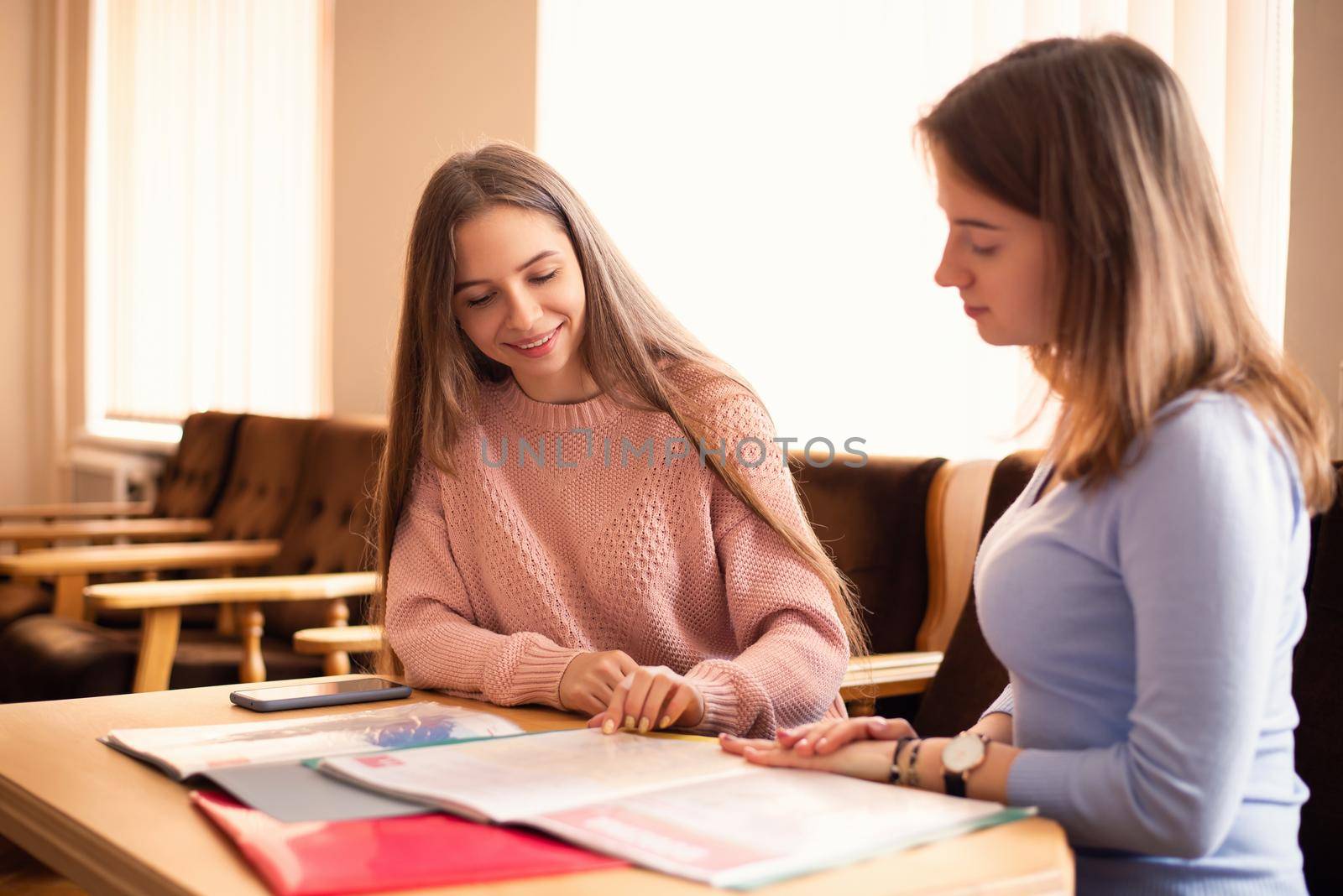 Two attractive female students sitting in the recreation room, preparing for the next class, communicating. Students life