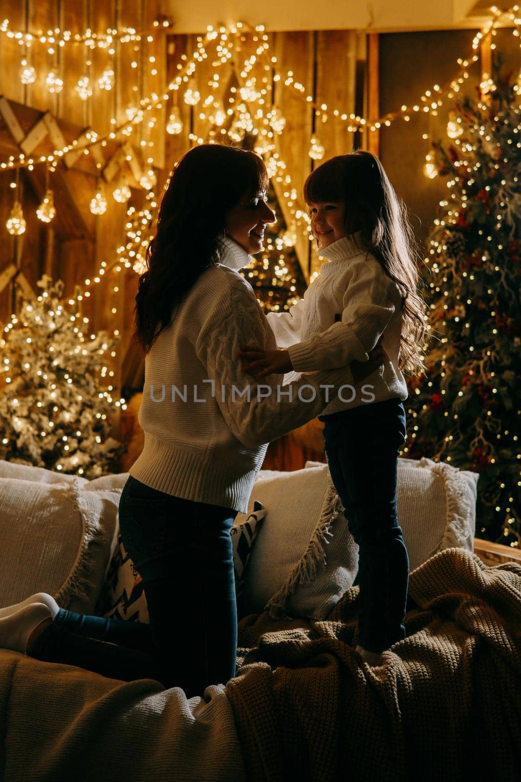 A little girl with her mother in a cozy home environment on the sofa next to the Christmas tree. The theme of New Year holidays and festive interior with garlands and light bulbs