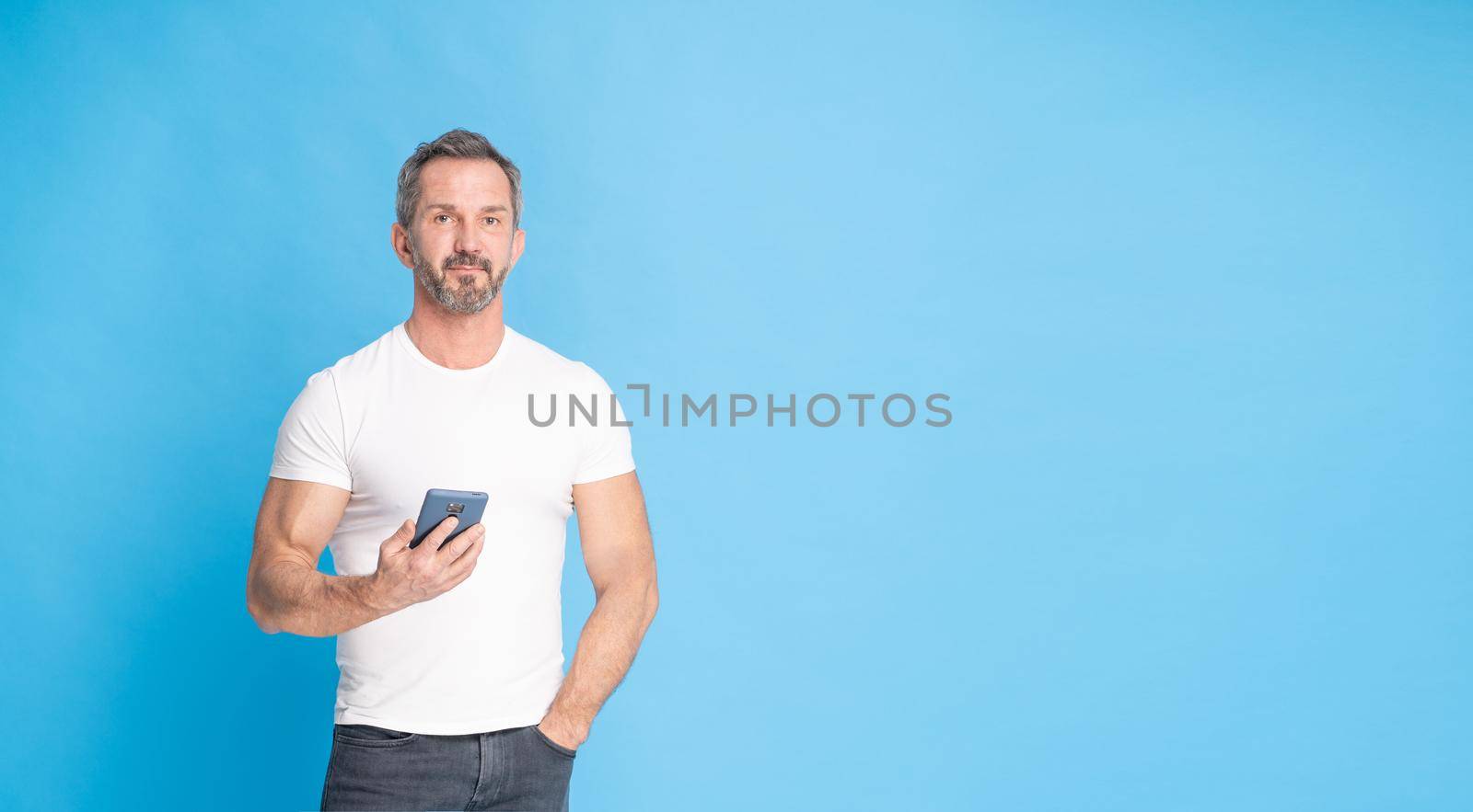 Handsome middle aged grey haired man 40s standing with with smartphone in hand wearing white t-shirt isolated on blue background. Mature fit man, healthy lifestyle concept. Copy space on right.