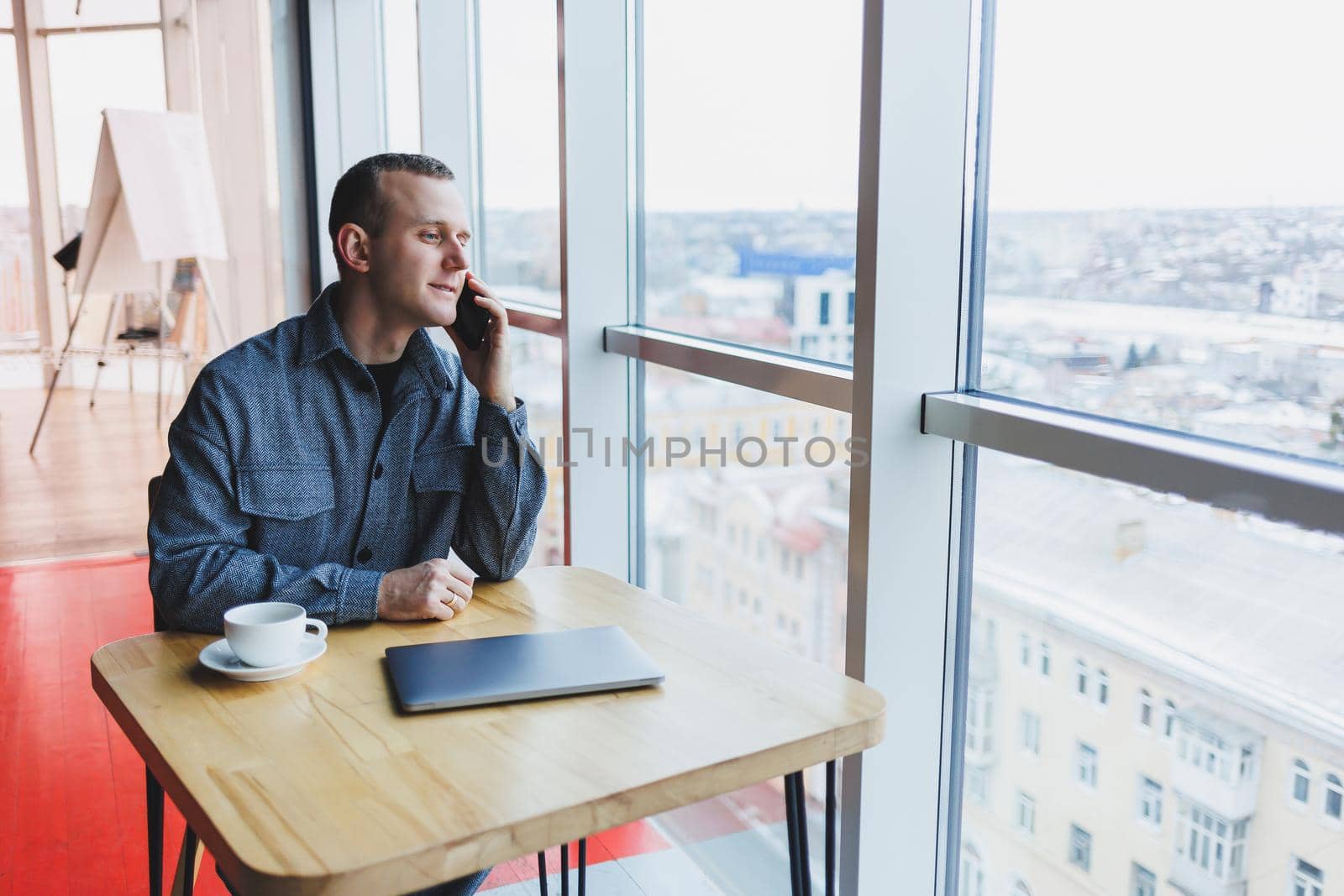 Happy man with coffee calling via smartphone while remote working laptop at work desk, cheerful freelancer enjoying coffee break while working remotely