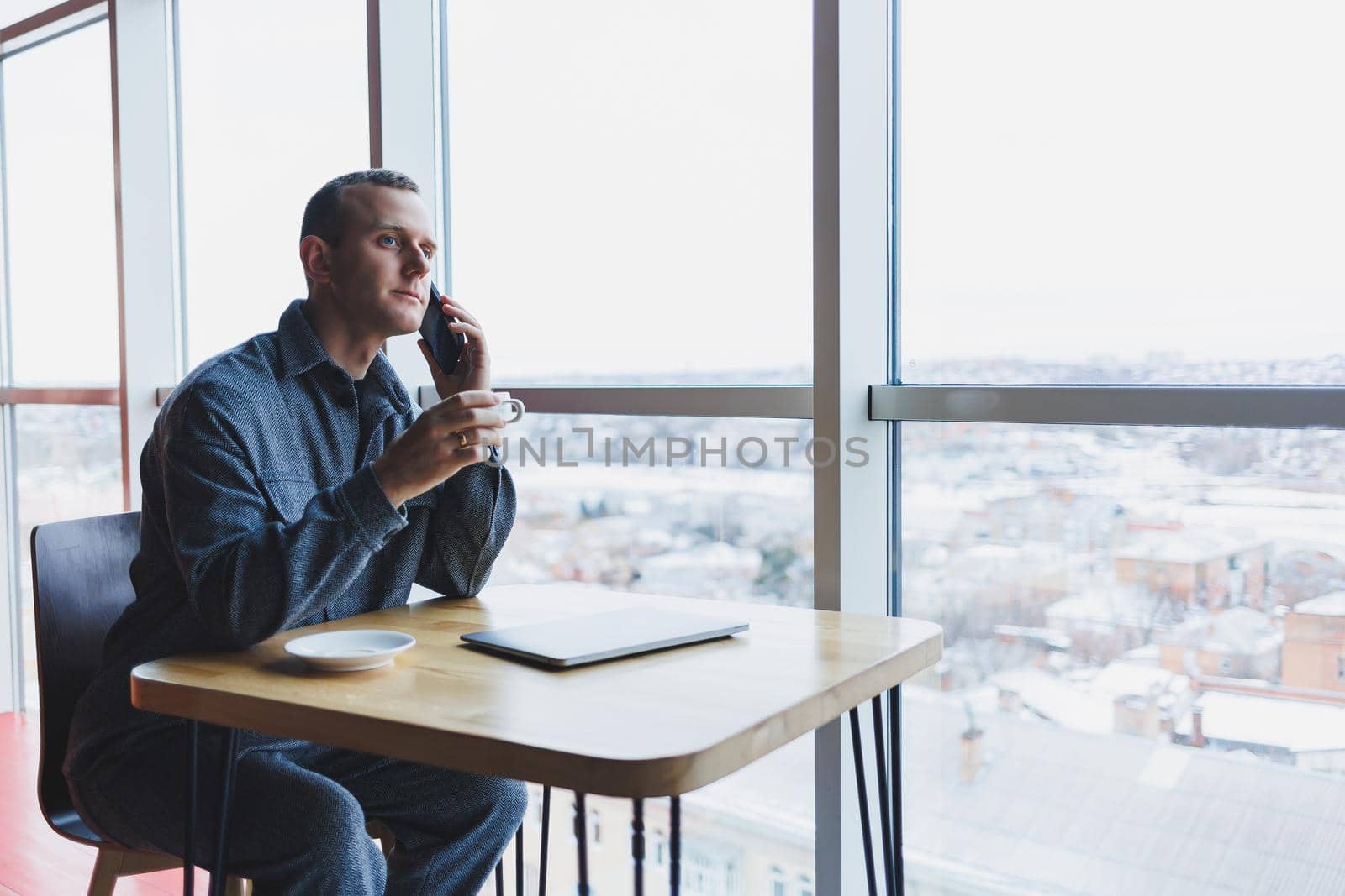 Happy man with coffee calling via smartphone while remote working laptop at work desk, cheerful freelancer enjoying coffee break while working remotely