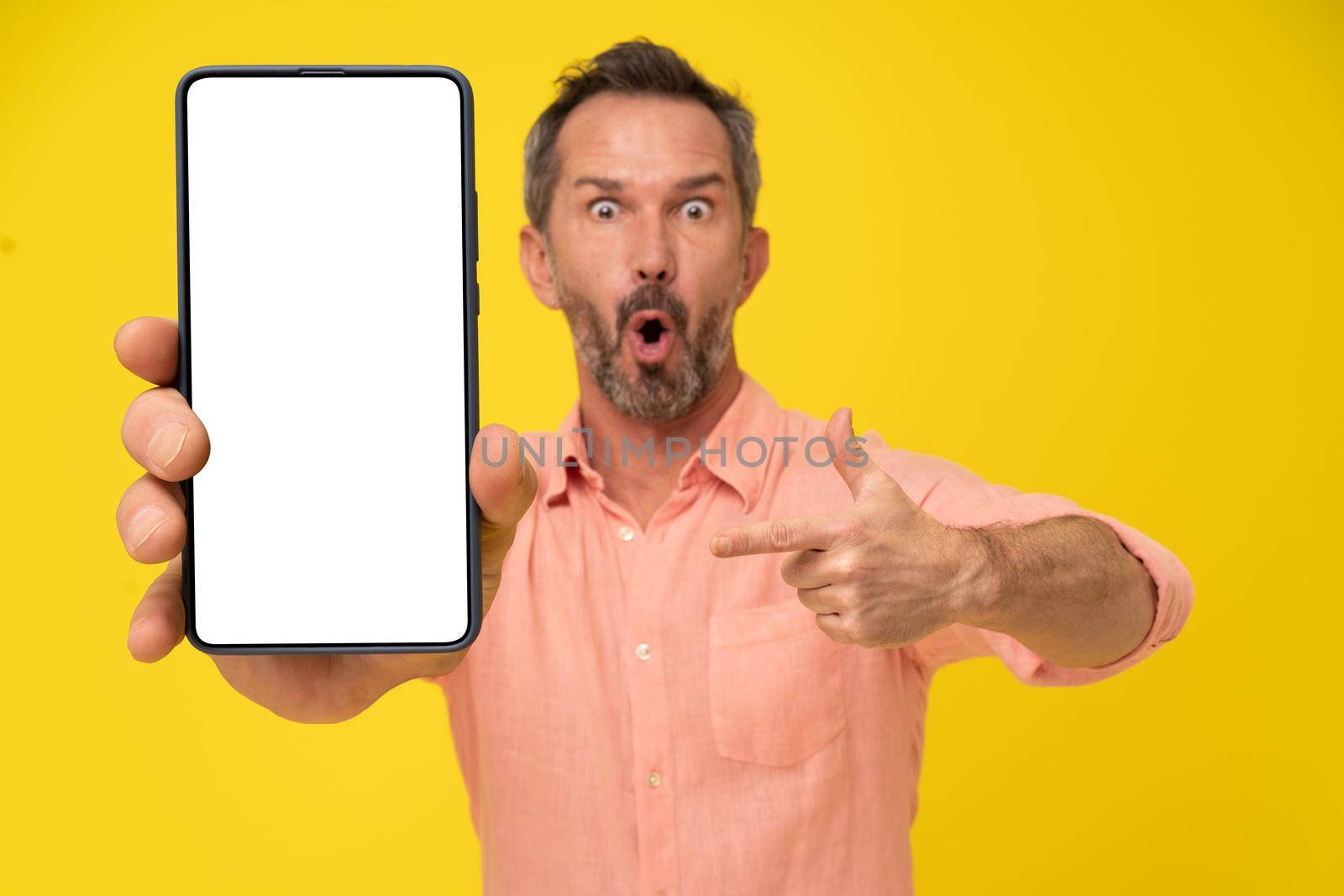 Excited, shocked of a new mobile app middle aged grey haired man pointing at smartphone with white screen in hand wearing peach shirt isolated on yellow. Mobile app advertisement by LipikStockMedia